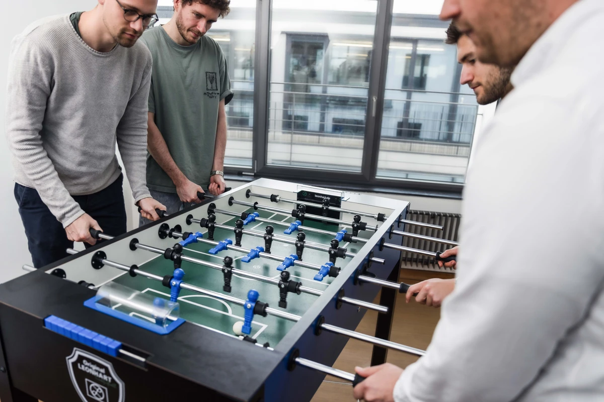 Vier Kollegen spielen im Pausenbereich im Büro am Tischkicker ein Match, bei dem die Spielergebnisse datenbasiert erfasst werden. Teamgeist und Transparenz beim gemeinsamen Kicker Match